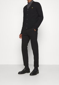 Schwarze Strickjacke mit V-Ausschnitt, fünf Knöpfen und einem gelben Emblem, kombiniert mit schwarzen Hosen und schwarzen Lederschuhen.