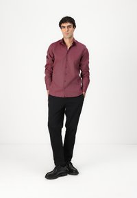 Chemise bordeaux à manches longues en tissu texturé, design à boutons, associée à un pantalon noir et des chaussures noires, se tenant devant un fond blanc.