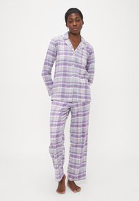 Žena stojí naboso v levanduľovo-bielej kockovanej pyžamovej súprave s dlhými rukávmi a nohavicami na bielom pozadí.