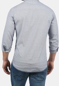 Uomo con camicia grigia chiaro a maniche lunghe, risvoltate, e jeans blu, in piedi di spalle su sfondo bianco.