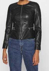 Giacca in pelle nera dal design aderente, con dettagli trapuntati e cerniere argentate davanti e sulle tasche. Abbinata a jeans in denim blu.