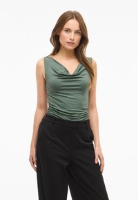 Top verde sin mangas con escote drapeado y textura fruncida, combinado con pantalones negros de tiro alto con bolsillos laterales.