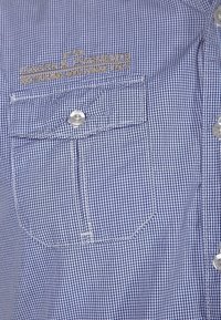 Camicia a quadri blu e bianca con taschino chiuso da un bottone, caratterizzata da testo ricamato in oro. Texture liscia con dettagli di cuciture bianche.