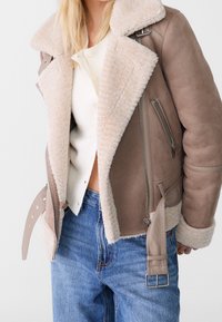 Bruine suède shearling jas met een beige pluche kraag, zijzakken en zilveren ritsdetails, gecombineerd met een crèmekleurige top en blauwe spijkerbroeken.