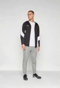 Zwart-witte sportjas met capuchon, gecombineerd met grijze joggingsbroek en zwarte schoenen. Glad materiaal, nauwsluitend ontwerp en logoakcenten.