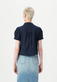 Mørkeblå kortermet bluse med puffede ermer, blonder rundt kragen og en rynket ryggdesign. Kombinert med et lyst denimskjørt.