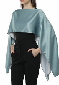 Satin grøn kappe med en kort design, der har en glat overflade og en kontrasterende hvid underside, båret over en sort top og bukser.