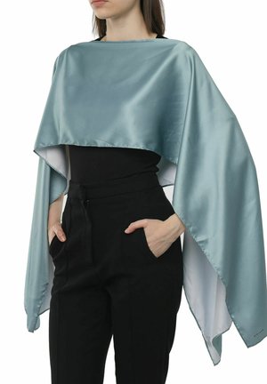 Satin grøn kappe med en kort design, der har en glat overflade og en kontrasterende hvid underside, båret over en sort top og bukser.