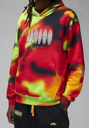 Person iført en multifarvet rød, gul og grøn Nike Jordan hættetrøje med sorte Nike Jumpman bukser, stående med hænderne ned langs siderne.