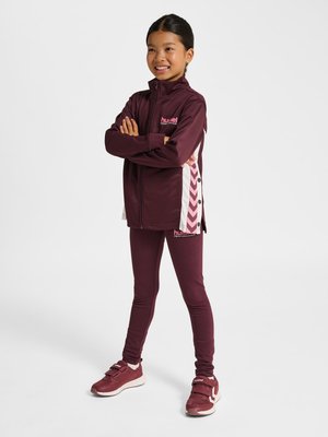 Meisje lacht met gekruiste armen, draagt een bordeaux sportjack en leggings met roze chevronstrepen, staand op een witte achtergrond.