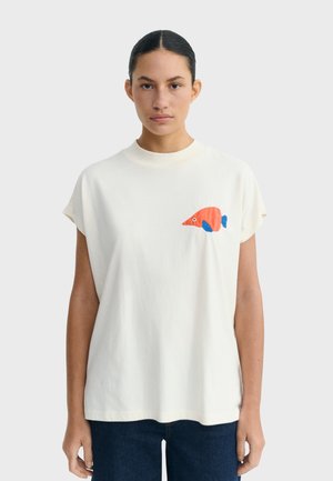 Kobieta o ciemnych włosach związanych w kok, ubrana w luźną białą koszulkę z małą grafiką pomarańczowo-niebieskiej ryby na piersi oraz ciemne spodnie.