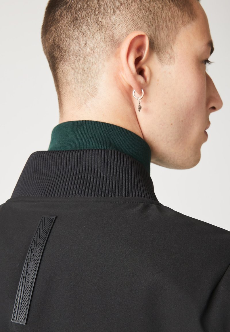 Veste noire avec col côtelé et doublure intérieure verte ; présente un panneau texturé noir au dos et une petite boucle d'oreille en argent.