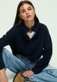 Navy blue knitted sweater with a high collar and button accents, paired with light blue distressed denim jeans and beige footwear.