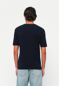 Mann med mørkt hår iført en enkel marineblå kortermet skjorte og lyseblå jeans, vendt bort fra kameraet mot en hvit bakgrunn.
