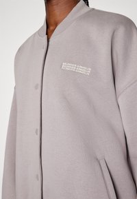 Felpa grigia oversize con una texture liscia, chiusura a bottoni a pressione e piccolo logo bianco "Under Armour" sul petto.