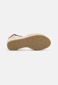 Beige espadrille korkokenkä, jossa on teksturoitu kangaspinta, ruskea nilkkahihna ja kumipohja, jossa on kohokuvioidut brändityyppiset yksityiskohdat.