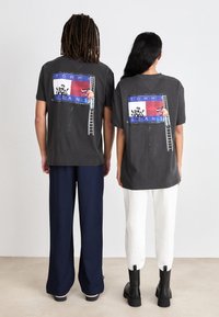 Camisetas gráficas grises con un estampado colorido en la parte trasera que muestra una escalera y figuras, combinadas con pantalones y botas en contraste.