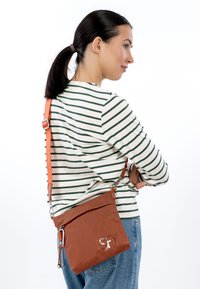 Brūna, teksturēta pleca soma ar regulējamu oranžu siksnu, aprīkota ar rāvējslēdzēja aizdari un izšūtu detaļu priekšpusē.