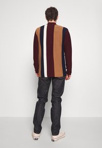 Suéter de punto con rayas verticales en burdeos, crema, azul y marrón. Combinado con jeans oscuros y zapatillas blancas. Vista trasera.