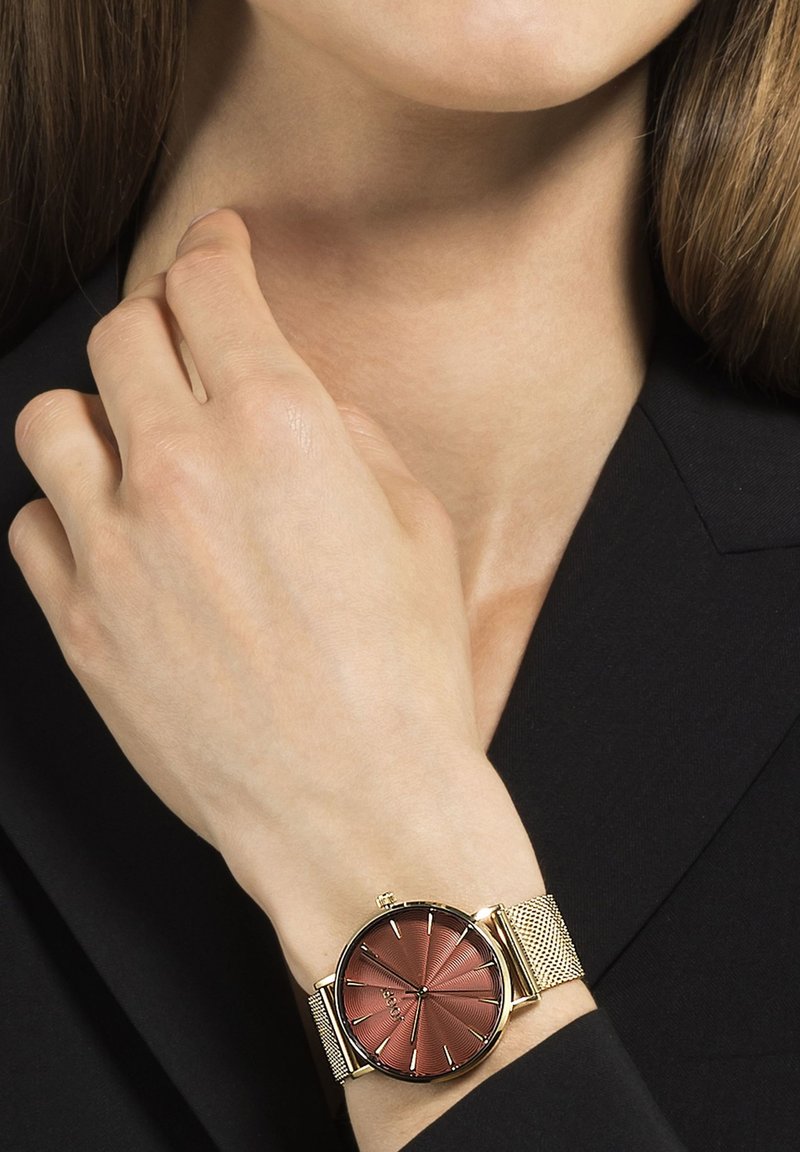 Runde Uhr mit einem burgunderfarbenen strukturierten Zifferblatt, goldenen Stundenmarkierungen und einem goldenen Mesh-Armband. Auf einem Handgelenk präsentiert, teilweise von Fingern verdeckt.