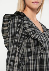 Blouse à carreaux noir et beige avec des manches à volants et un décolleté en V. Le tissu est léger avec une finition texturée et un accent superposé.