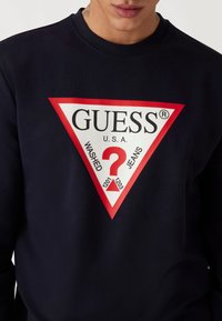 Felpa navy con un grande logo triangolare rosso e bianco che presenta "GUESS," "WASHED," e un punto interrogativo, realizzata in morbido cotone.