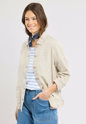 Femme souriante, portant une veste beige boutonnée sur une chemise rayée bleu et blanc, un jean bleu, et un foulard marine à rayures blanches autour du cou.