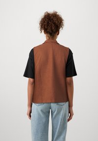 Bruine mouwloze vest met een gestructureerde afwerking, gecombineerd met een zwart T-shirt met korte mouwen en lichtblauwe jeans, gezien van de achterkant.