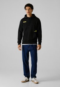 Sudadera negra con bolsillo canguro, acentos en amarillo en el logo y capucha con cordón, combinada con jeans de mezclilla azules y zapatillas blancas.