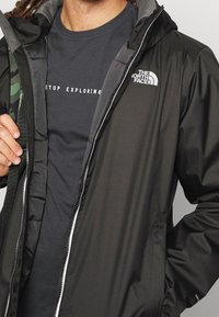 Persona vistiendo una chaqueta negra de North Face sobre una camiseta oscura que dice "Deja de explorar", sujetando la correa de una mochila de camuflaje con una mano.