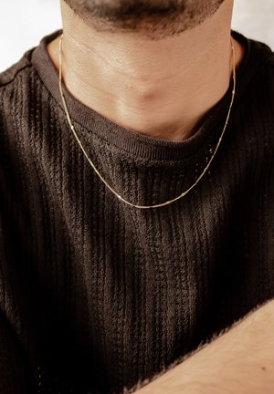 Goldene Kettenhalskette, die über einem strukturierten dunkelbraunen Pullover getragen wird, zeigt ein glattes, dünnes Design mit subtilen Details.