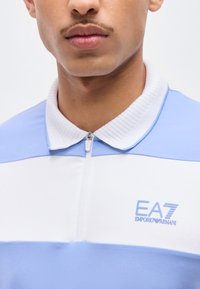 Κοντινό πλάνο ενός ατόμου που φορά μπλε και λευκή ρίγα πόλο μπλούζα με φερμουάρ και το λογότυπο EA7 Emporio Armani στο στήθος.