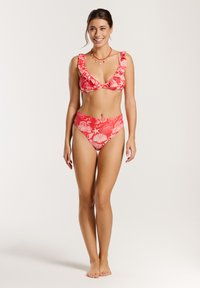 Piros bikini szett, fehér kagyló és tengeri csillag mintázattal. A melltartón fodros pántok és mély dekoltázs található; az alsó rész magas derékú.