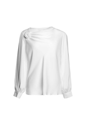 Blouse blanche à manches longues, col rond, poignets bouffants et fleur en tissu décorative sur l’épaule gauche.