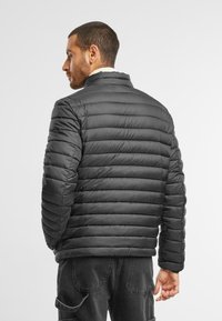 Sort puffjakke med en struktureret krave, vandret quiltet design og en glat, skinnende tekstur. Bagfra ses en tætsiddende silhuet.