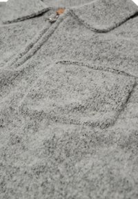 Szürke, texturált gyapjú ing, gallérral, gombos záródással és egyetlen rávarrt zsebbel az elején. Puha, bolyhos megjelenés.