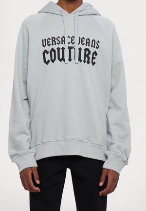 Persoon gekleed in een lichtgrijze hoodie met zwarte "Versace Jeans Couture" tekst, gecombineerd met een zwarte broek, gefotografeerd van nek tot heupen.