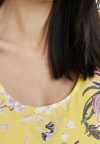 Keltainen kukkakuviollinen paita, jossa on pyöreä pääntie, violetit ja valkoiset kukkakuviot sekä pehmeä, sileä kangas.