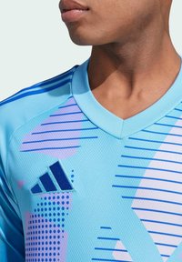 Grembiule sportivo azzurro chiaro con scollatura a V, caratterizzato da motivi geometrici in viola e blu, tessuto testurizzato e logo Adidas sul lato sinistro del petto.
