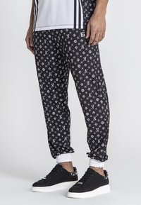 Zwarte joggers met een herhalend wit logopatroon, elastische boorden en zijzakken, gedragen met zwarte sneakers met witte zolen.