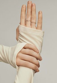 Top de manga larga crema con puños enrollados, manos adornadas con dos delicadas anillas de oro que presentan pequeñas piedras preciosas agrupadas, mostrando una textura suave.