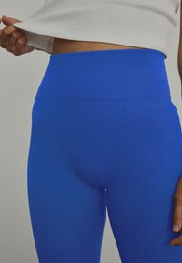 Leggings azules acanaladas con una cintura alta, que presentan una textura suave y un ajuste ceñido, mostrados con una camiseta blanca que se levanta ligeramente.