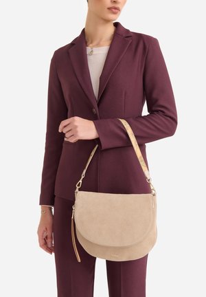 Donna con un completo bordeaux che tiene una borsa in suede beige con una tracolla decorata a motivo, appoggiata al braccio, su uno sfondo bianco semplice.