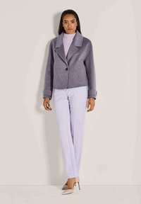 Frau steht, trägt eine lila Wolljacke, einen lavendelfarbenen Rollkragenpullover, helllila Hosen und beigefarbene High Heels.