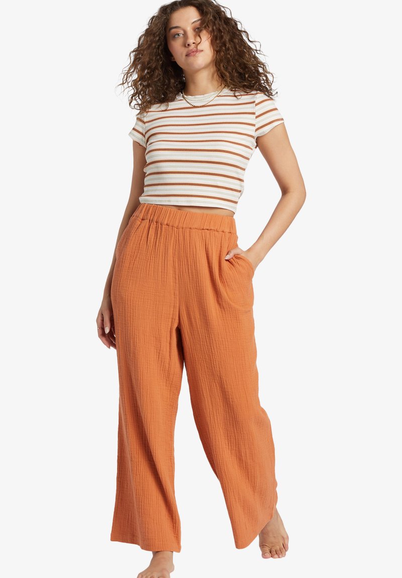 Striped, cropped t-shirt in white with orange hues, paired with loose-fitting, textured, rust-orange pants with an elastic waistband and pockets.