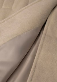 Beżowa tkanina polarowa o miękkiej fakturze, pokryta gładką podszewką w kolorze taupe, z kontrastującymi szwami i subtelnym połyskiem.