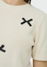 Crème-kleurige gebreide trui met korte mouwen en een ribbelkraag, versierd met twee zwarte lintstrikjes als accenten.
