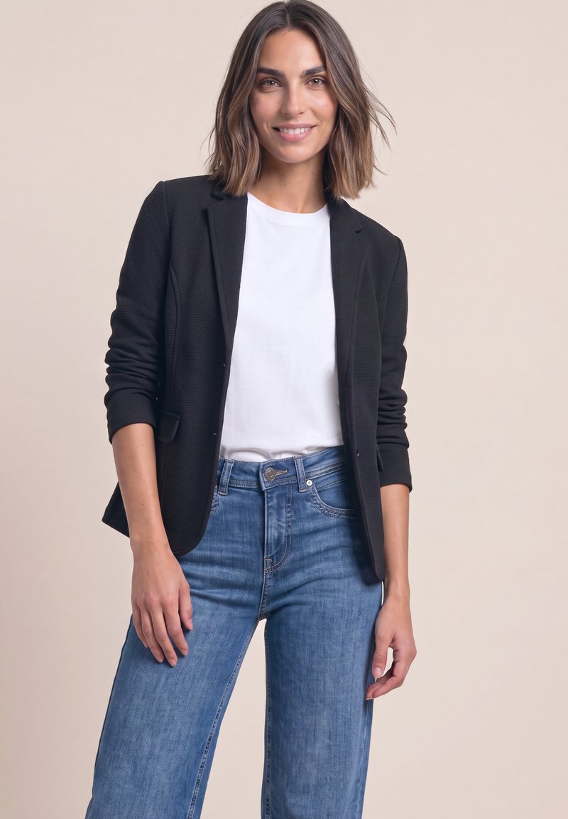 Jeune femme aux cheveux bruns mi-longs sourit, portant un blazer noir, une chemise blanche et un jean bleu sur un fond beige uni.