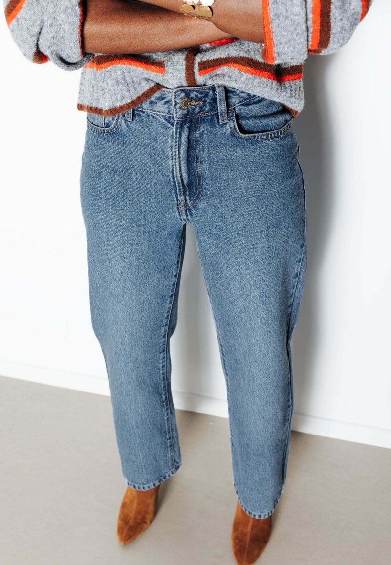 Personne portant un jean bleu taille haute à jambes droites, un pull gris avec des rayures orange et des bottines en daim marron à bouts pointus, les bras croisés.