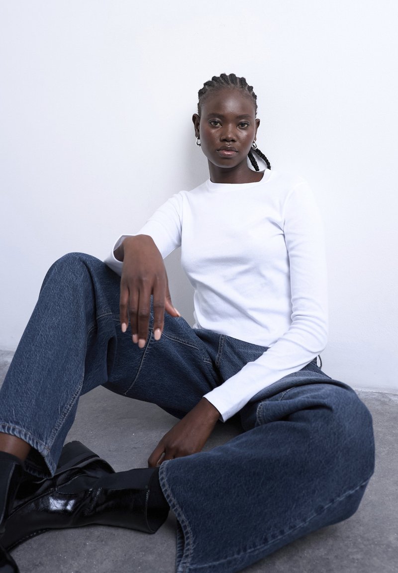 Νεαρή γυναίκα με πλεξούδες κάθε ται στο πάτωμα δίπλα σε λευκό τοίχο, φορώντας λευκό μακρυμάνικο πουκάμισο, τζιν με φαρδιά μπατζάκια και μαύρα μποτάκια.